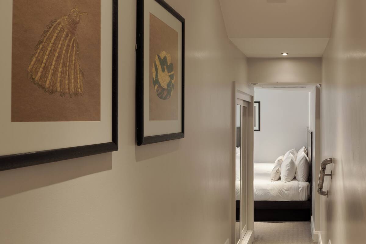 Un pasillo con obras de arte enmarcadas en las paredes, una cama con ropa de cama blanca al fondo y una puerta que da a otra habitación.
