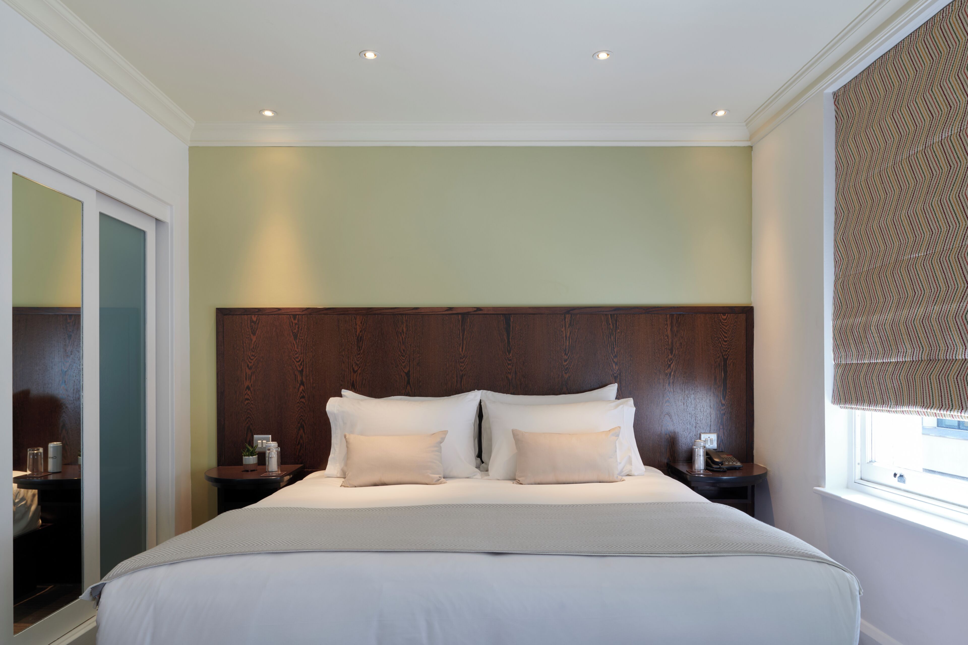 Une chambre d’hôtel avec un grand lit, deux tables de chevet, une tête de lit en bois et une fenêtre avec un store à motifs.