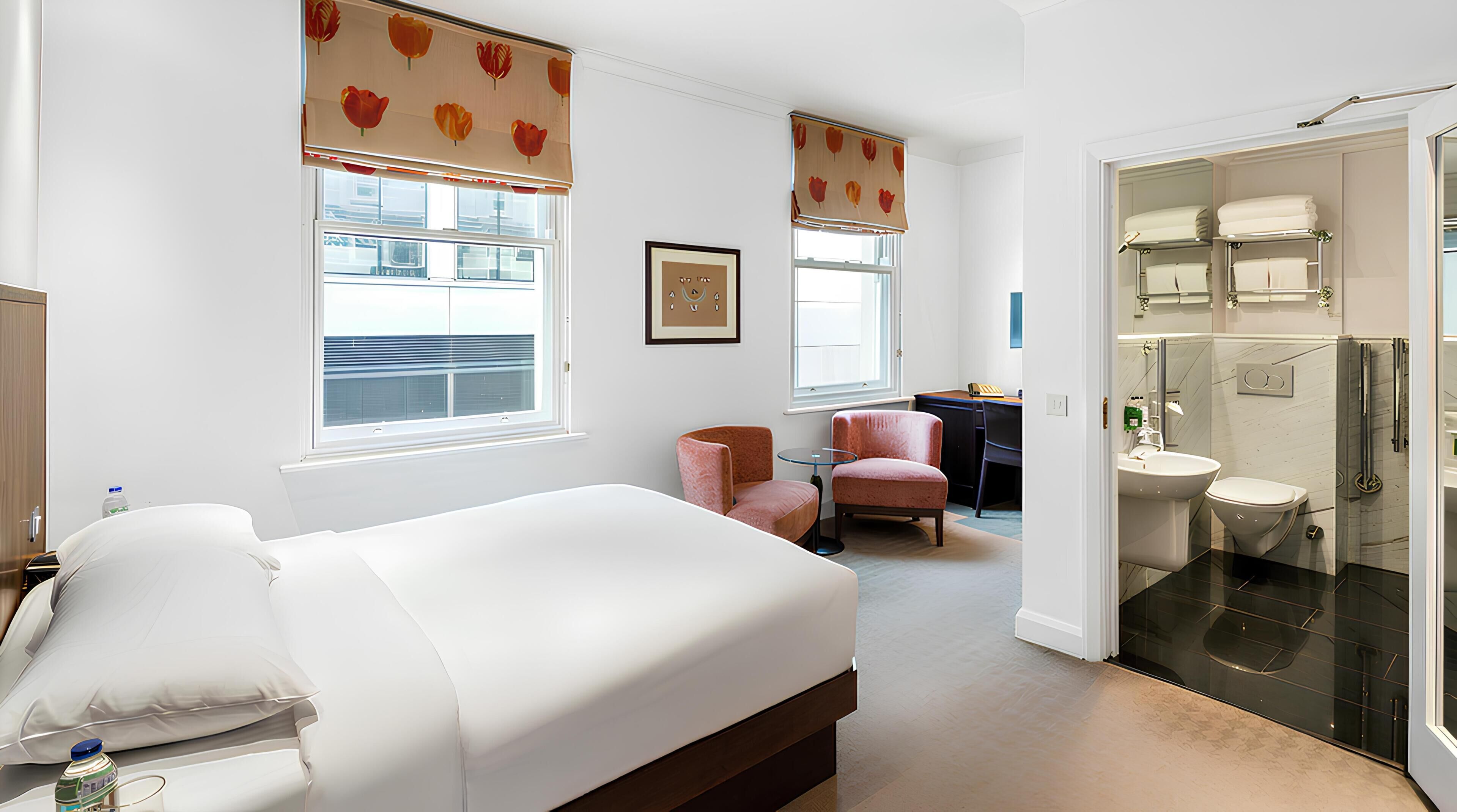A hotel room with a bed, a desk, a chair, a bathroom with a glass shower enclosure, and a view of the outside.