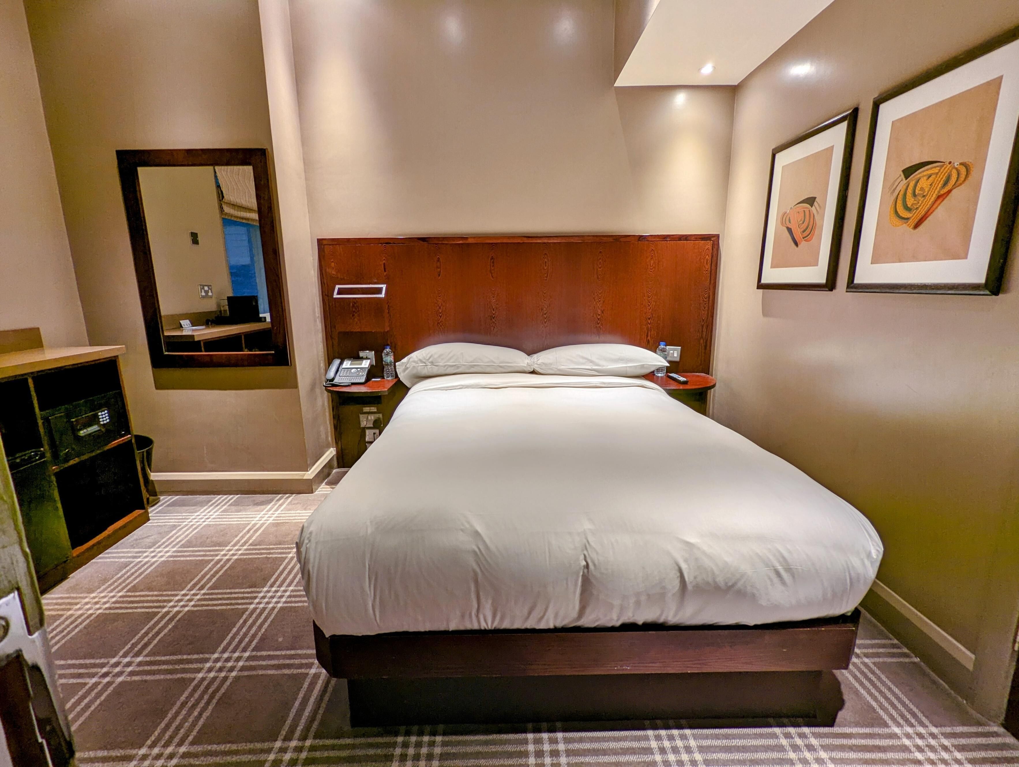 Une chambre d’hôtel avec un grand lit, une table de chevet avec un téléphone et une bouteille, un miroir et des œuvres d’art encadrées accrochées au mur.