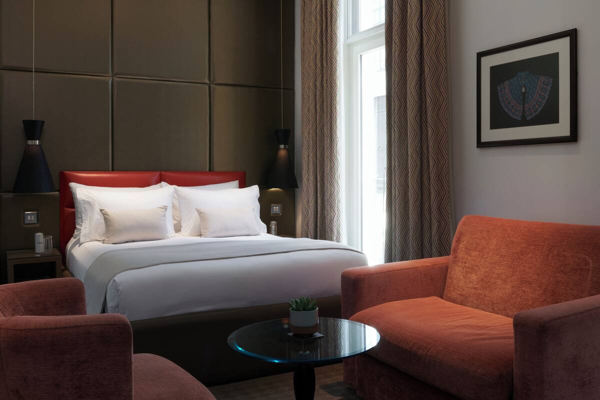 Habitación de hotel con una cama, dos sillones rojos, una mesita con una planta, un cuadro en la pared y una ventana con cortinas.