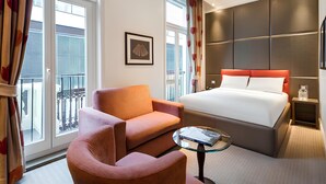 A hotel room with a bed, a chair, a small table, a framed picture, and large windows.