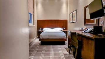 A hotel room with a bed, a desk, a chair, a television, and a bedside table with a lamp.