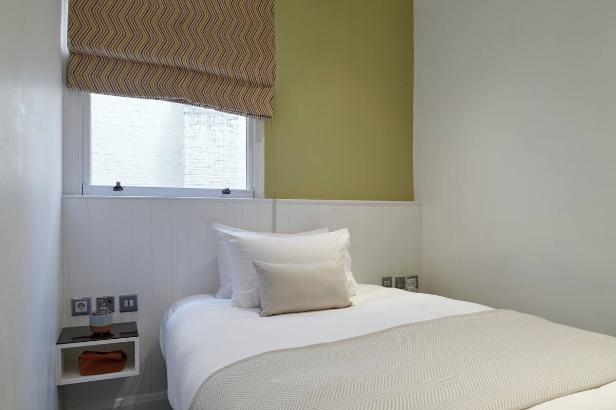 Habitación de hotel con cama, ventana con persianas, mesita de noche con lámpara y toma de corriente en la pared.
