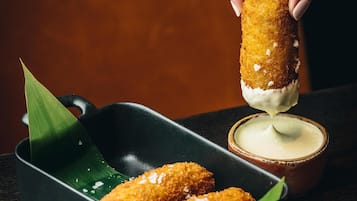 A hand dipping a breaded item into a creamy sauce, with a bowl of sauce and a dish of similar items on a wooden table.