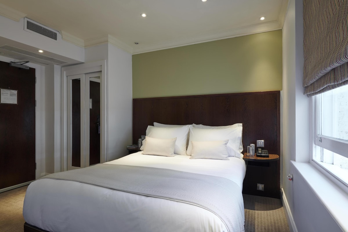 A hotel room with a large bed, a nightstand, a window with curtains, a door, and a vent on the ceiling.