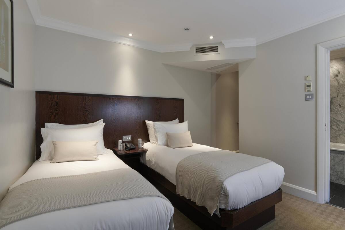 Habitación de hotel con dos camas, cabecero de madera, mesita de noche con teléfono y puerta de baño.