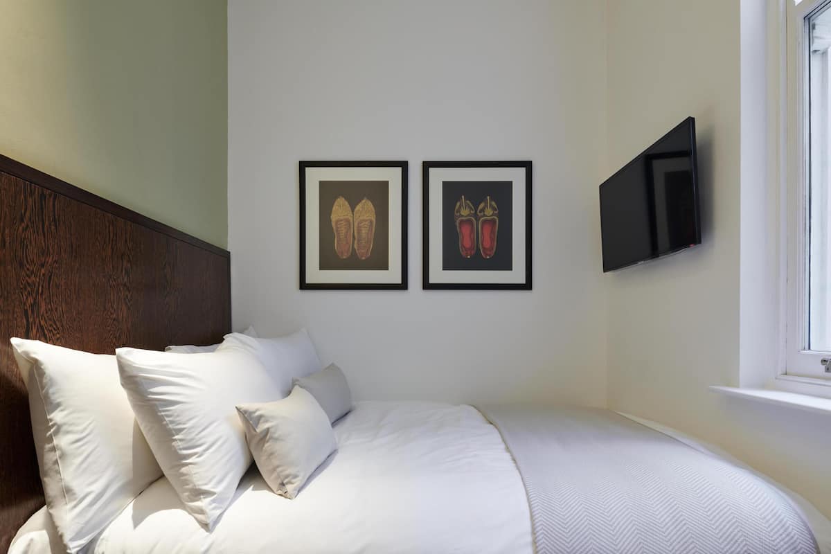 Una cama bien tendida con ropa blanca, dos cuadros en la pared y un televisor montado.