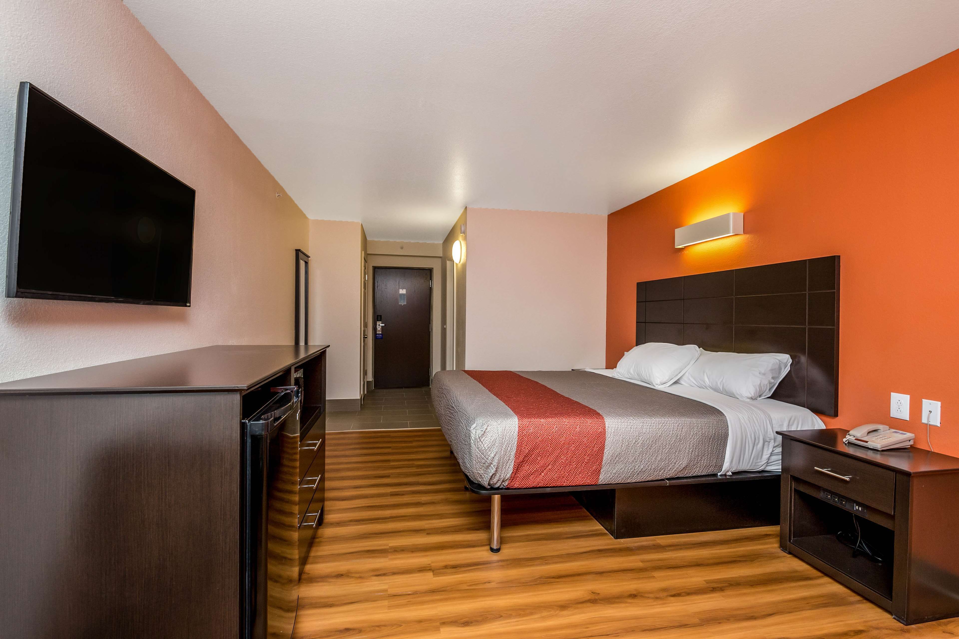 Een moderne hotelkamer met een grote flatscreen-tv, een bed met een rood-grijze plaid, een houten vloer en een klein bureau met een telefoon.