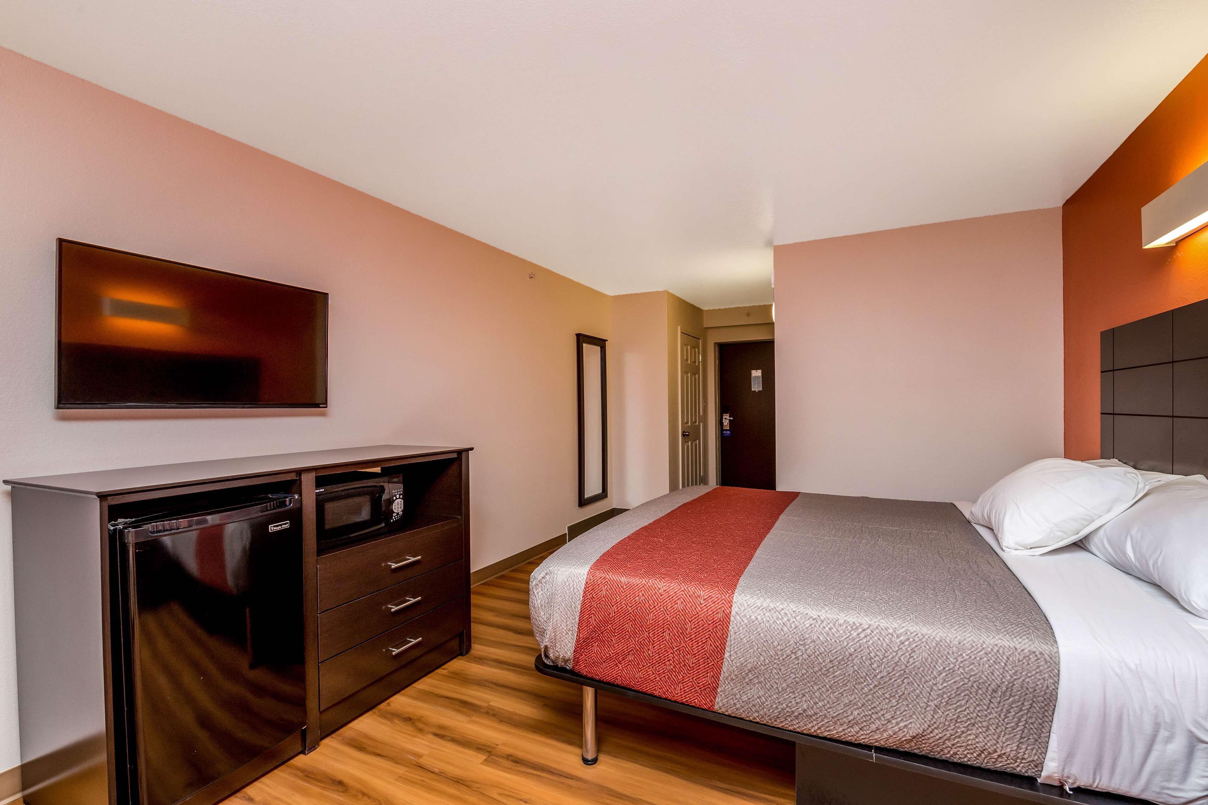 Quarto de hotel moderno com uma cama, penteadeira, televisão e porta para outro espaço.