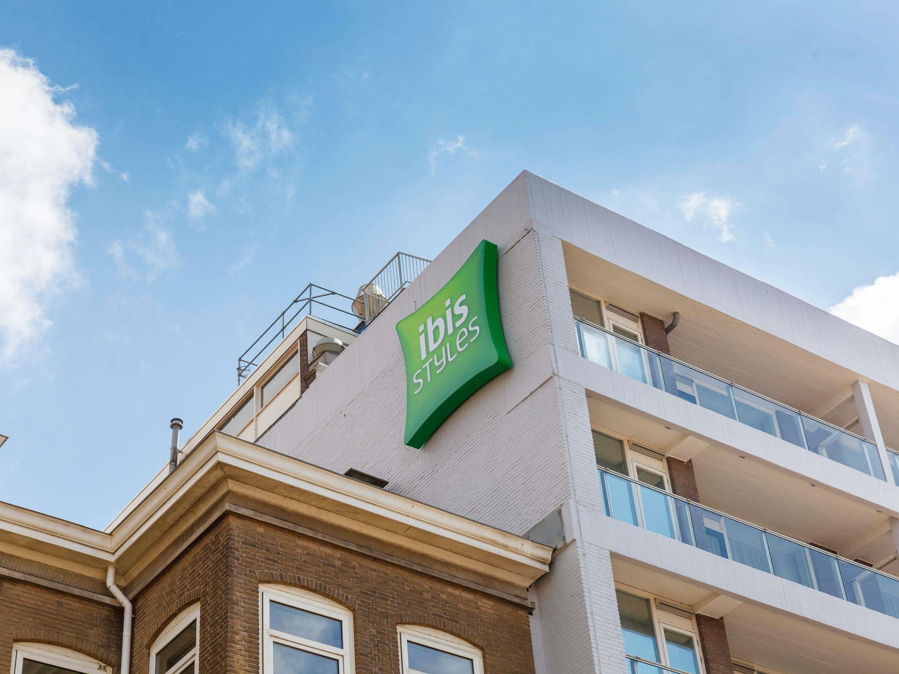 A building with a green 'ibis Styles' sign, a brick facade, and a modern balcony.