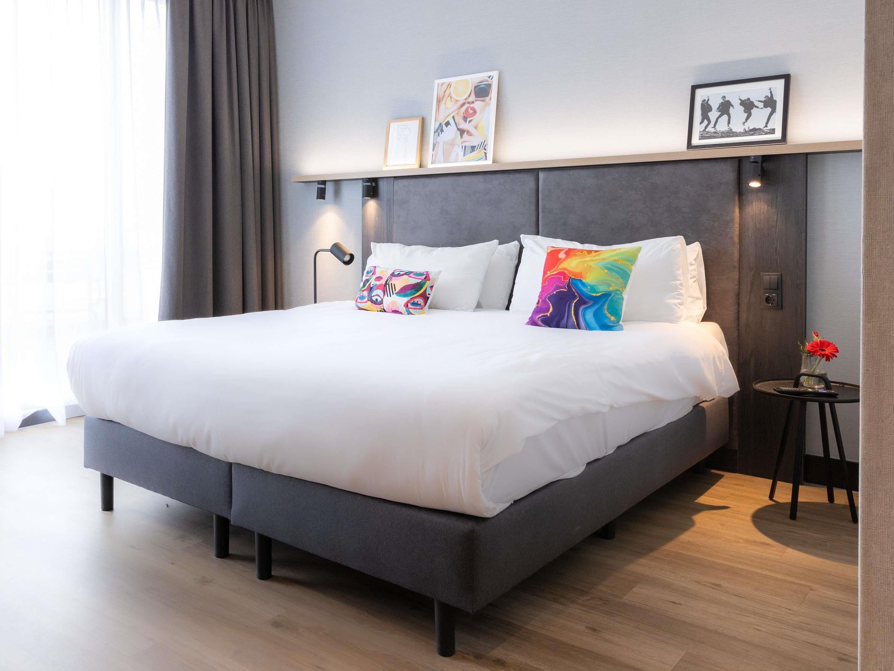 A modern hotel room with a large bed, a bedside table with a vase of flowers, and a wall-mounted shelf with framed pictures.