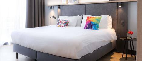Una habitación de hotel moderna con una cama grande, una mesita de noche con un jarrón de flores y un estante con marcos.
