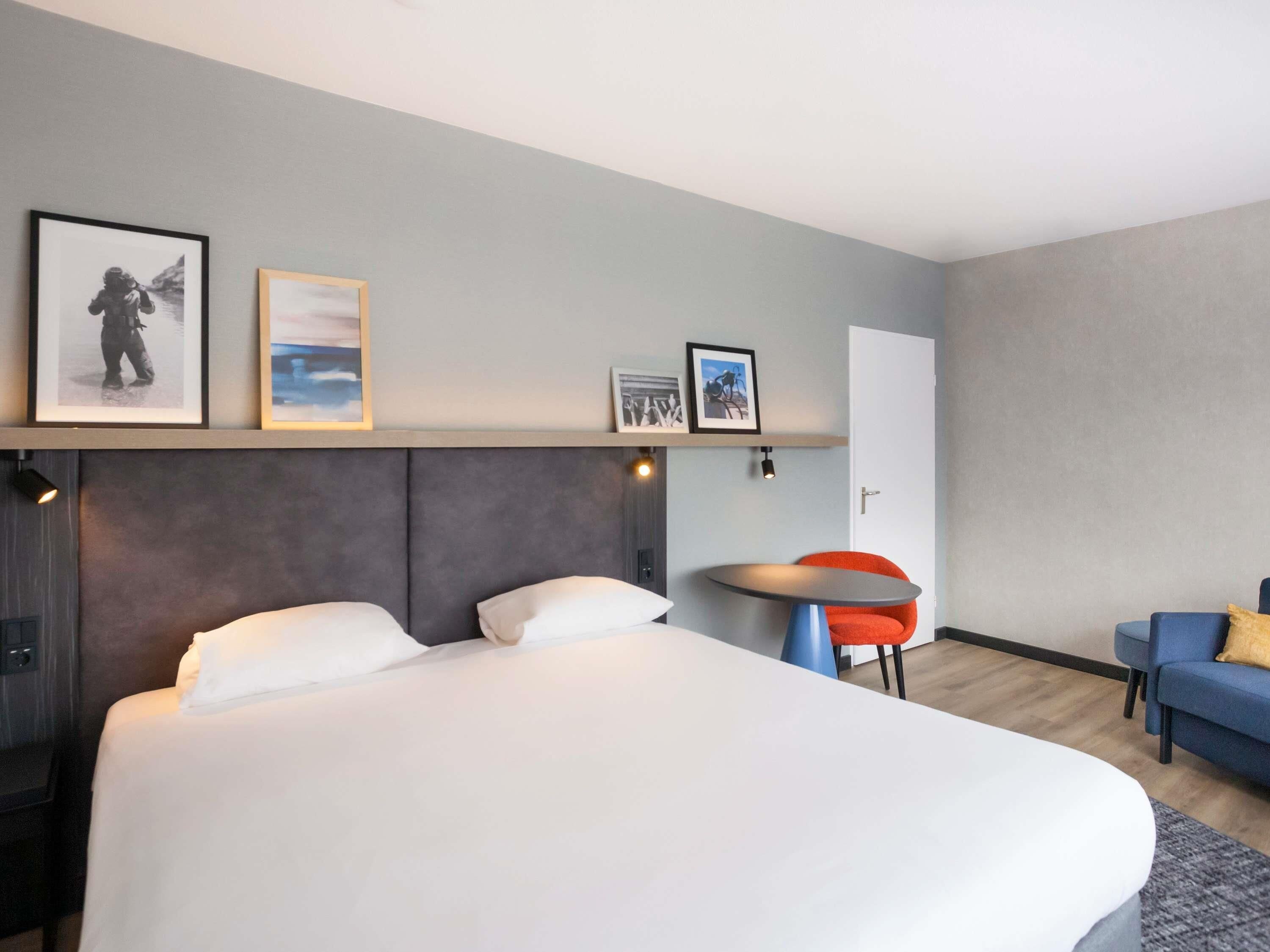 A modern hotel room with a large bed, a small round table, a blue armchair, and a red chair.