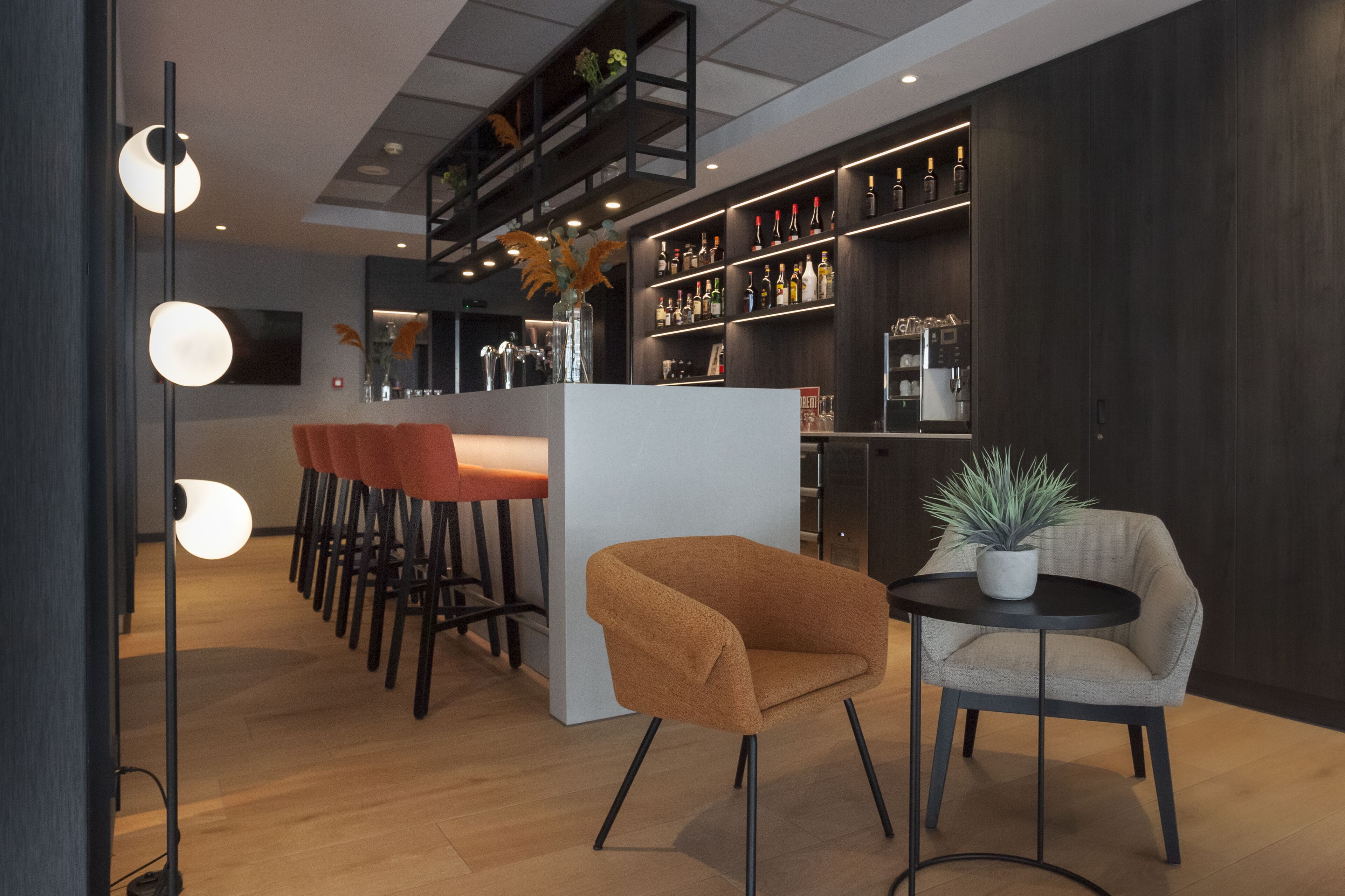 A modern bar area with a white counter, multiple bar stools, a round table with a potted plant, and a dark wooden shelving unit with bottles.