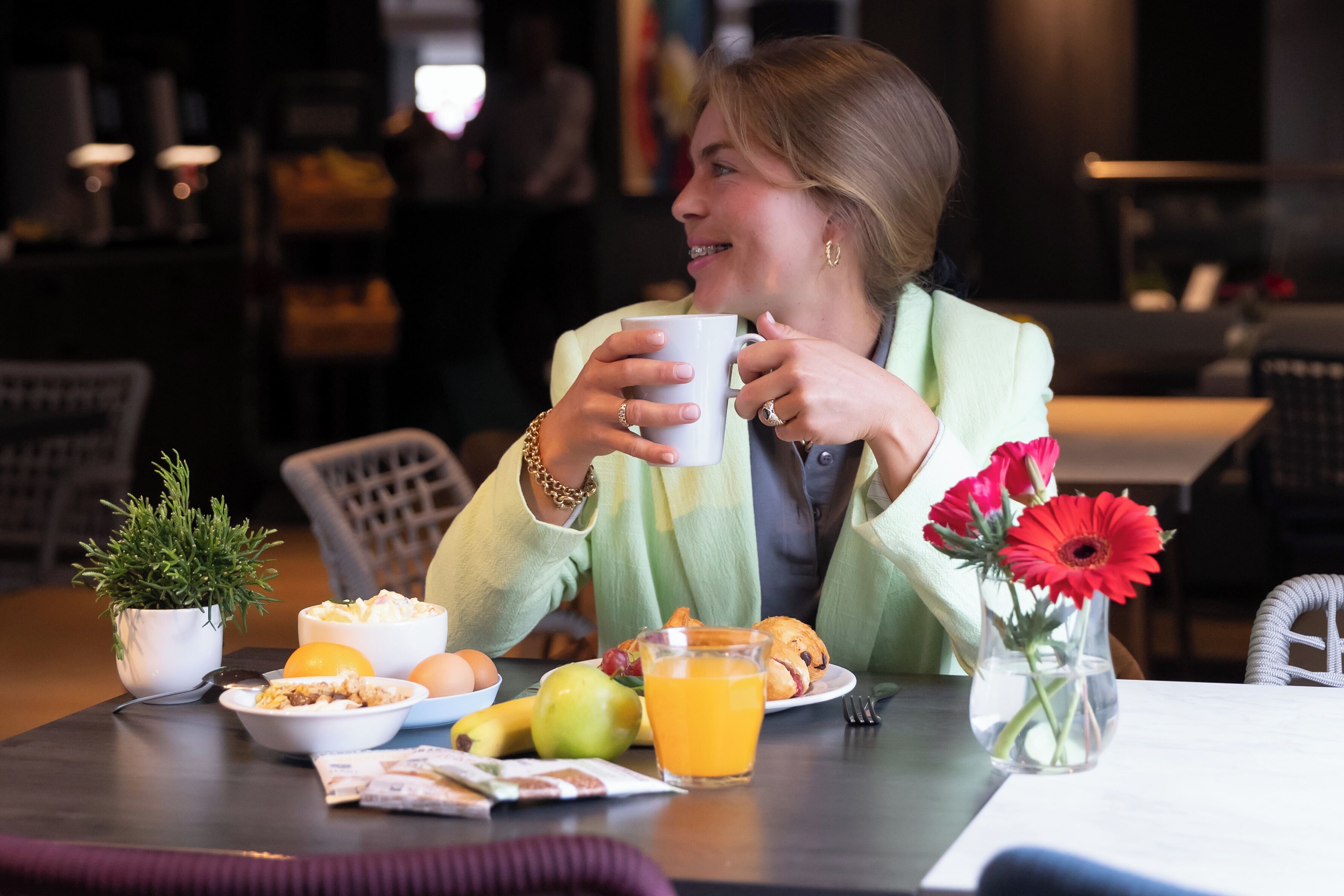 A person sitting at a table with breakfast items including eggs, fruit, and a drink. There is a small plant and a vase with flowers on the table.
