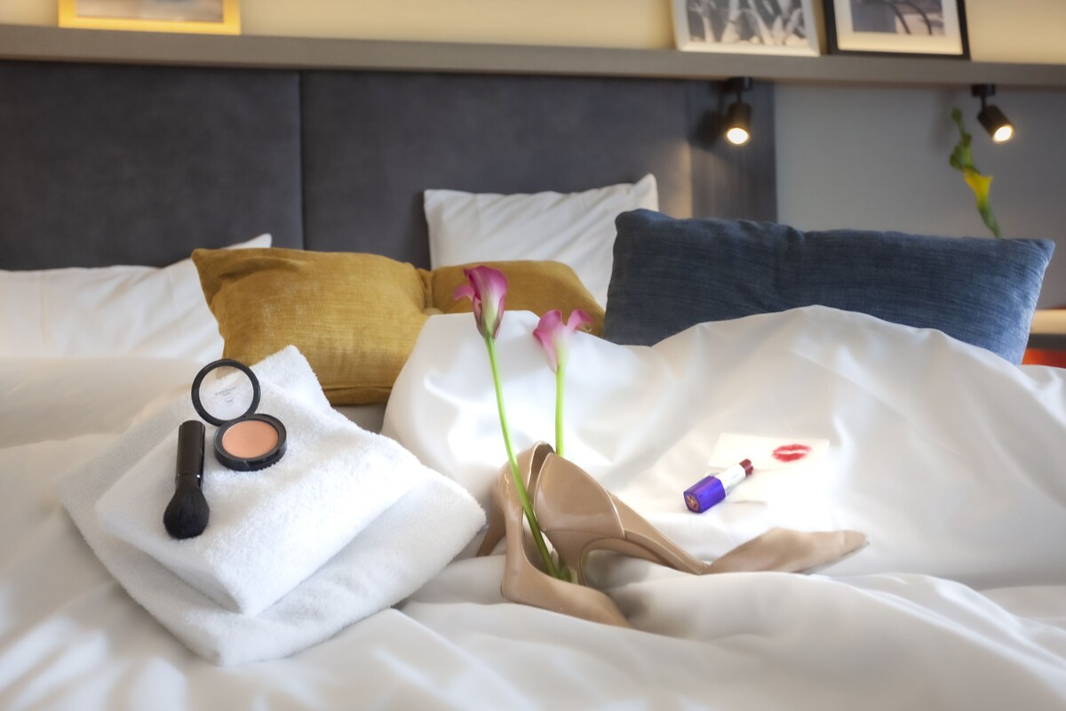 A neatly made bed with pillows, a makeup brush, a compact, a pair of shoes, and a vase with flowers.
