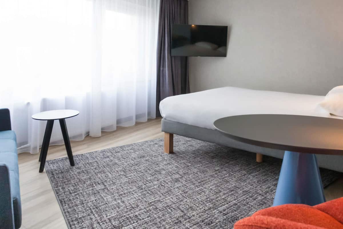 A modern hotel room with a bed, a TV mounted on the wall, a small round table, and a blue chair.