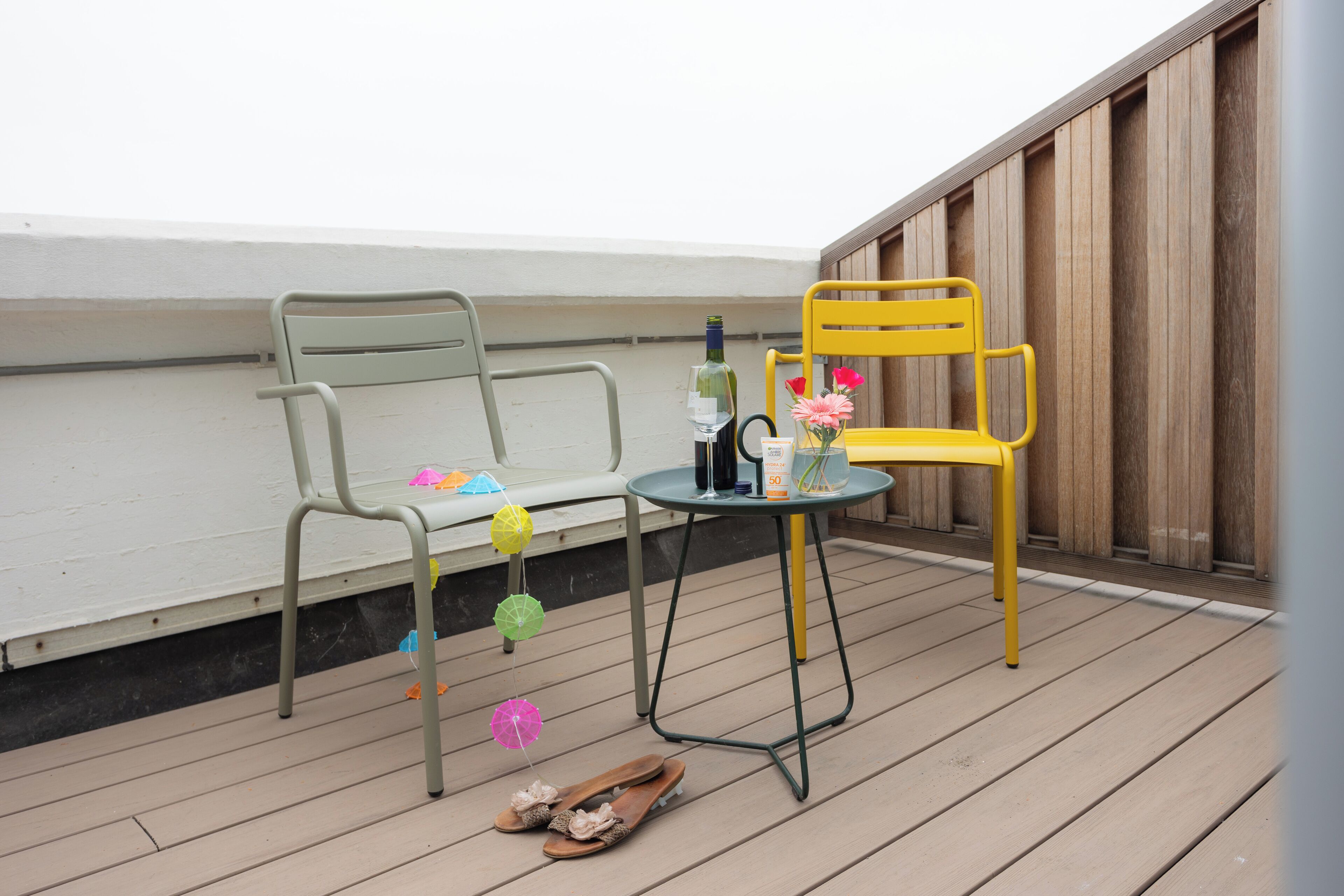 A wooden deck with a green metal bench and a yellow chair, a small round table with a bottle and flowers, and a pair of sandals.