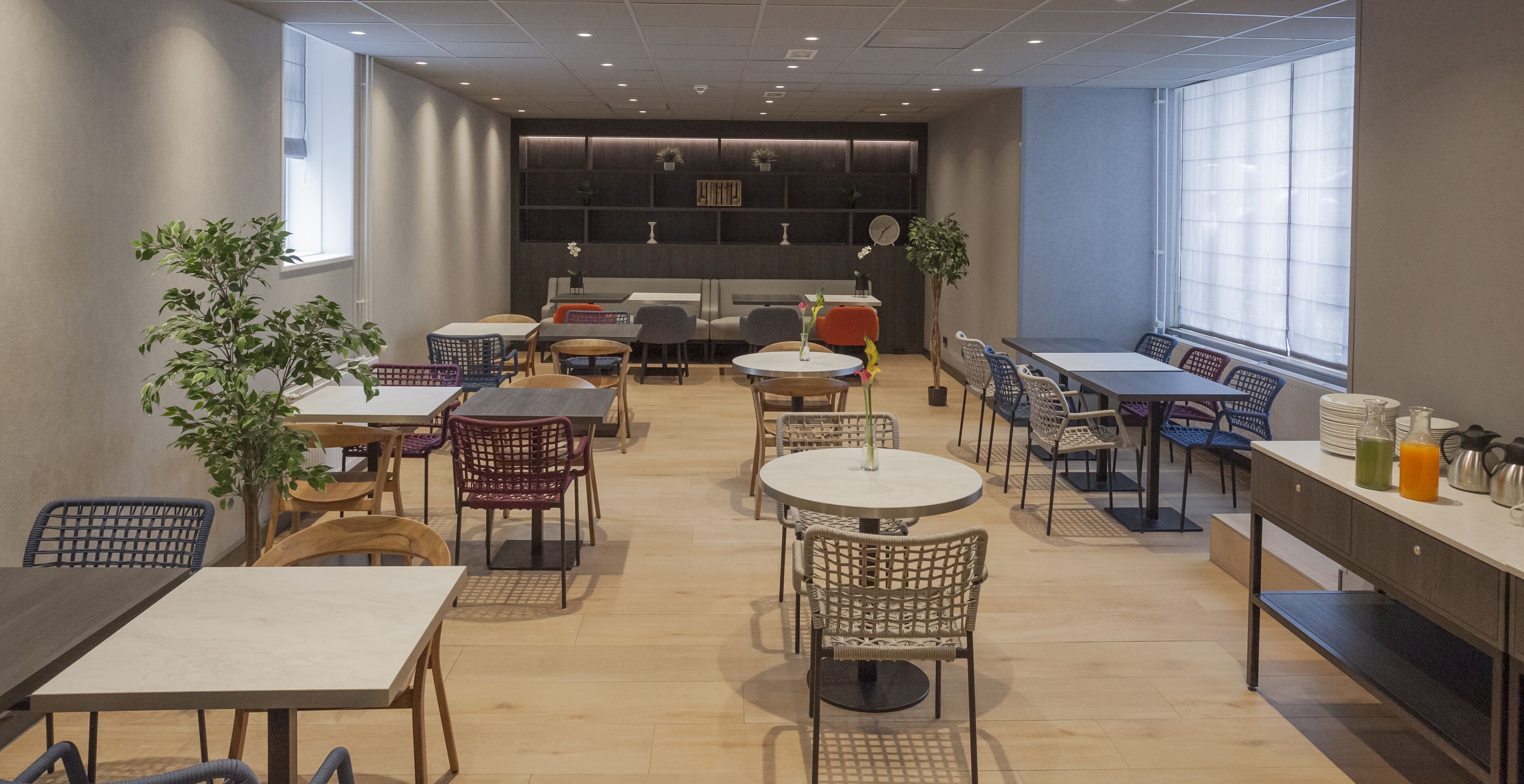 A modern dining area with multiple tables and chairs, a kitchen counter with breakfast items, and large windows providing natural light.