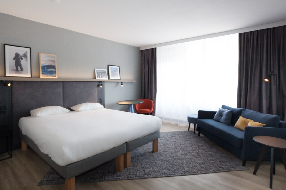 A modern hotel room with a large bed, a sofa, a small table, and a chair. There are framed pictures on the wall and a window with curtains.