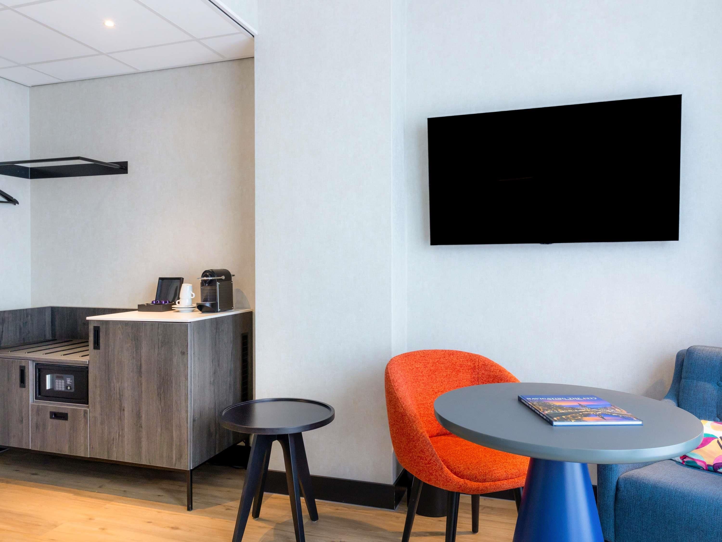A modern hotel room with a kitchenette, a flat-screen TV, a round table, and an orange chair.