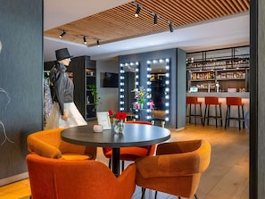 A modern interior with a round table, orange chairs, a bar area with stools, and a mannequin in a black outfit. - ibis Styles Den Haag Scheveningen (The Hague)