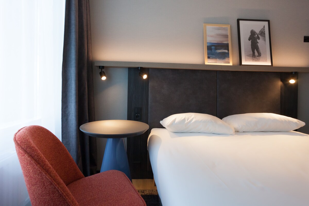 A hotel room with a bed, a red chair, a small round table, and two framed pictures on the wall.