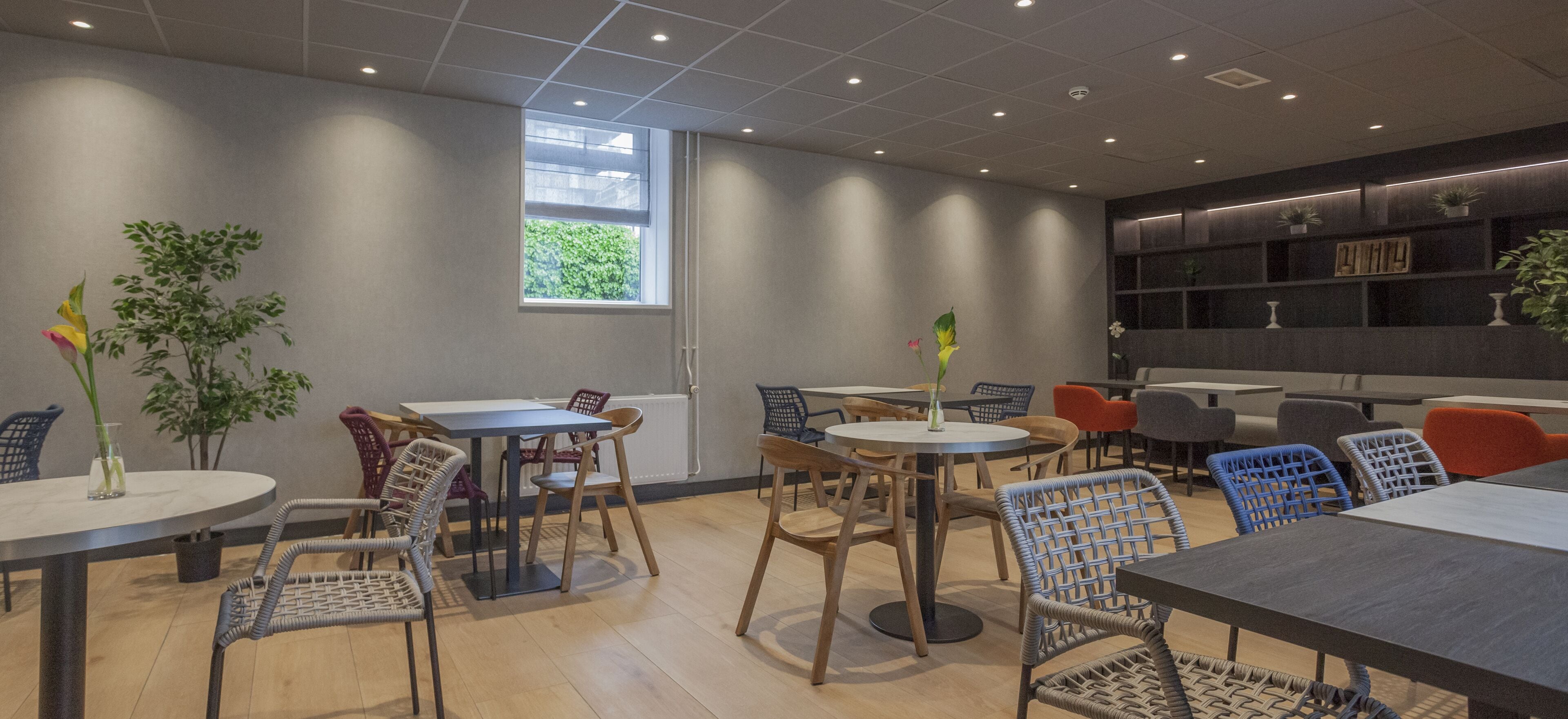 A modern interior with round tables and wicker chairs, a large window, and a bookshelf with plants.