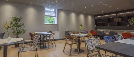 A modern interior with round tables and wicker chairs, a large window, and a bookshelf with plants.