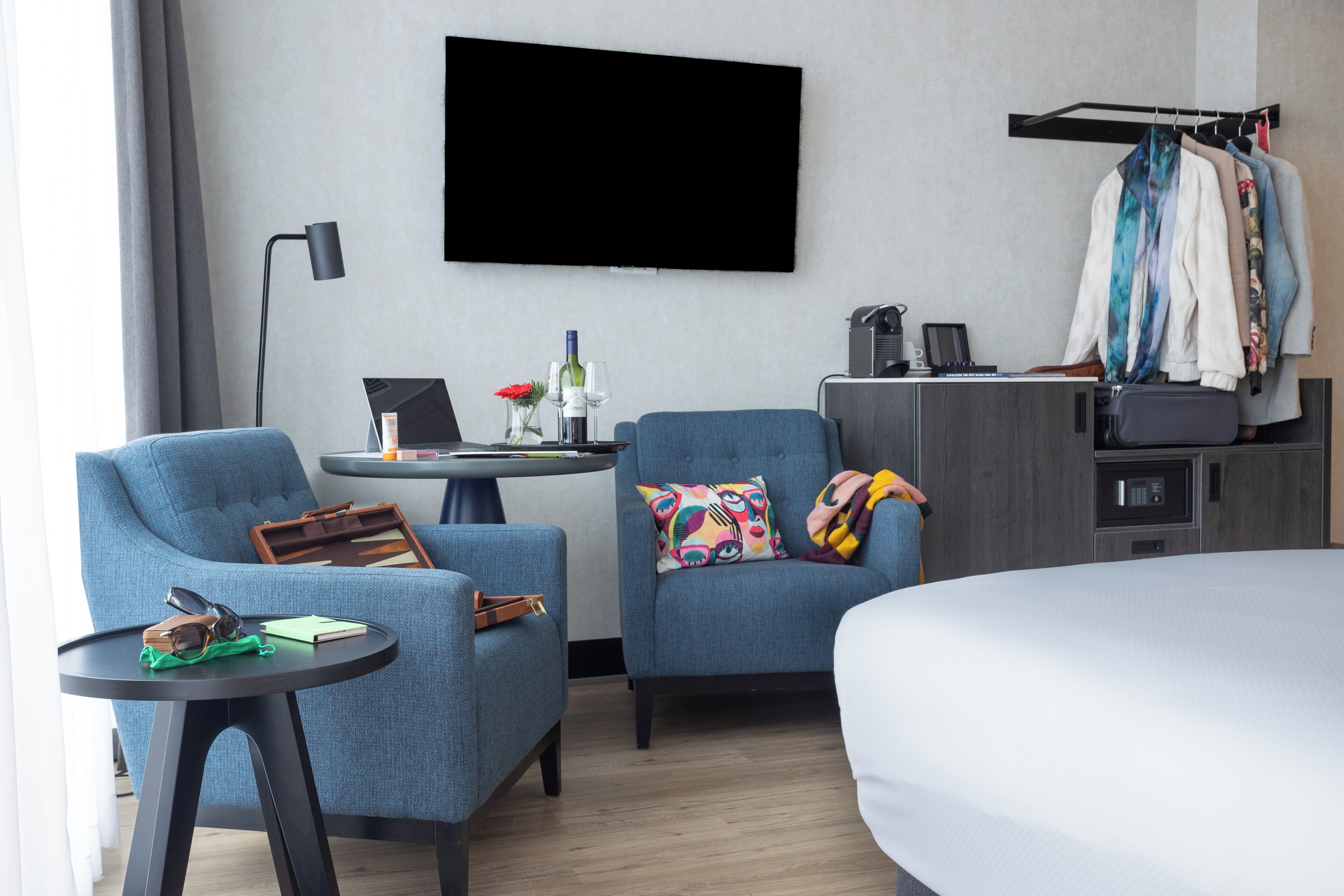 Una habitación de hotel con cama, dos sillones, una mesita con una computadora portátil, televisión montada en la pared, perchero con ropa y un microondas.