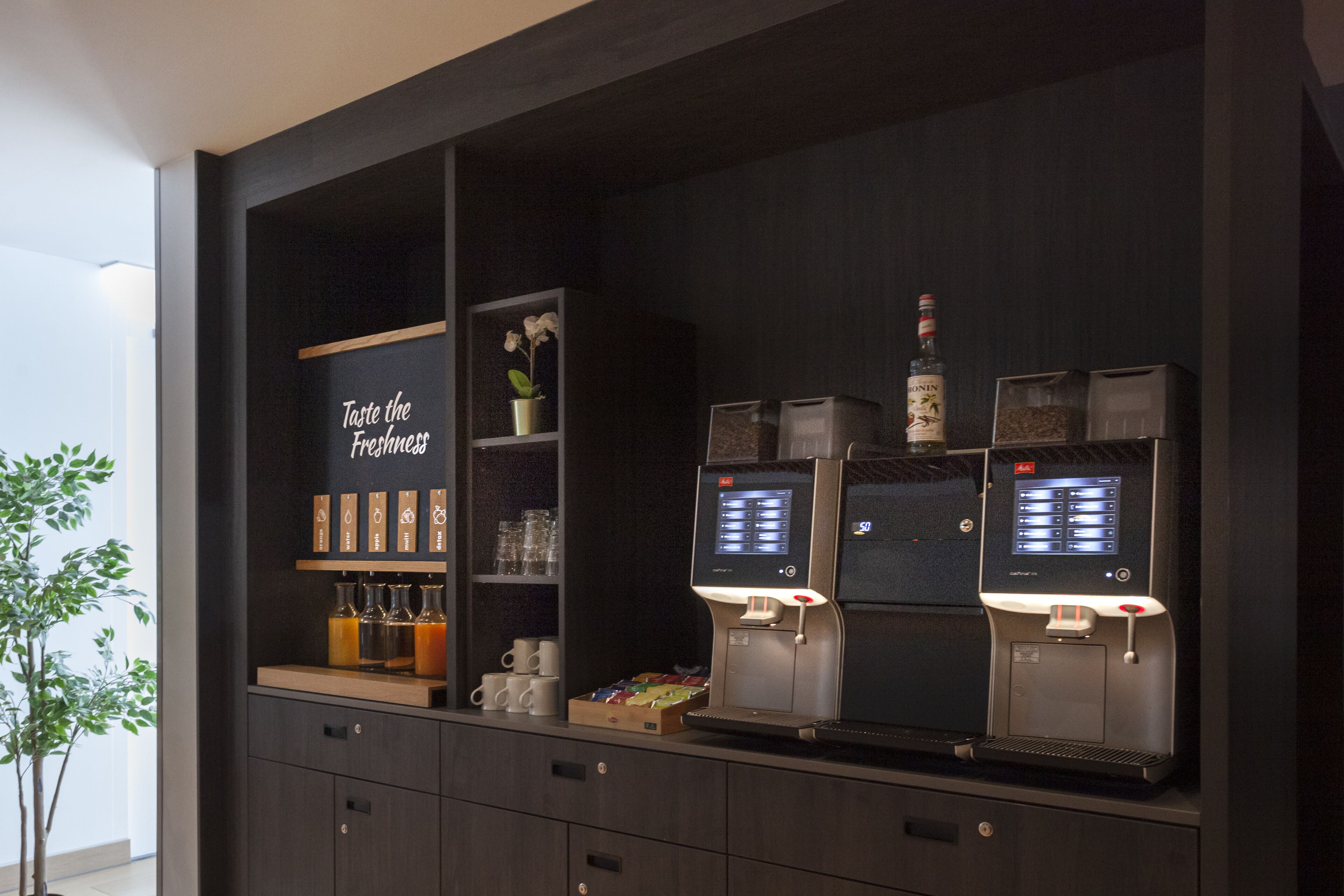Eine Barbereich mit zwei Espressomaschinen, einer Auswahl an Getränken und einem Schild mit der Aufschrift 'Probieren Sie die Frische'.