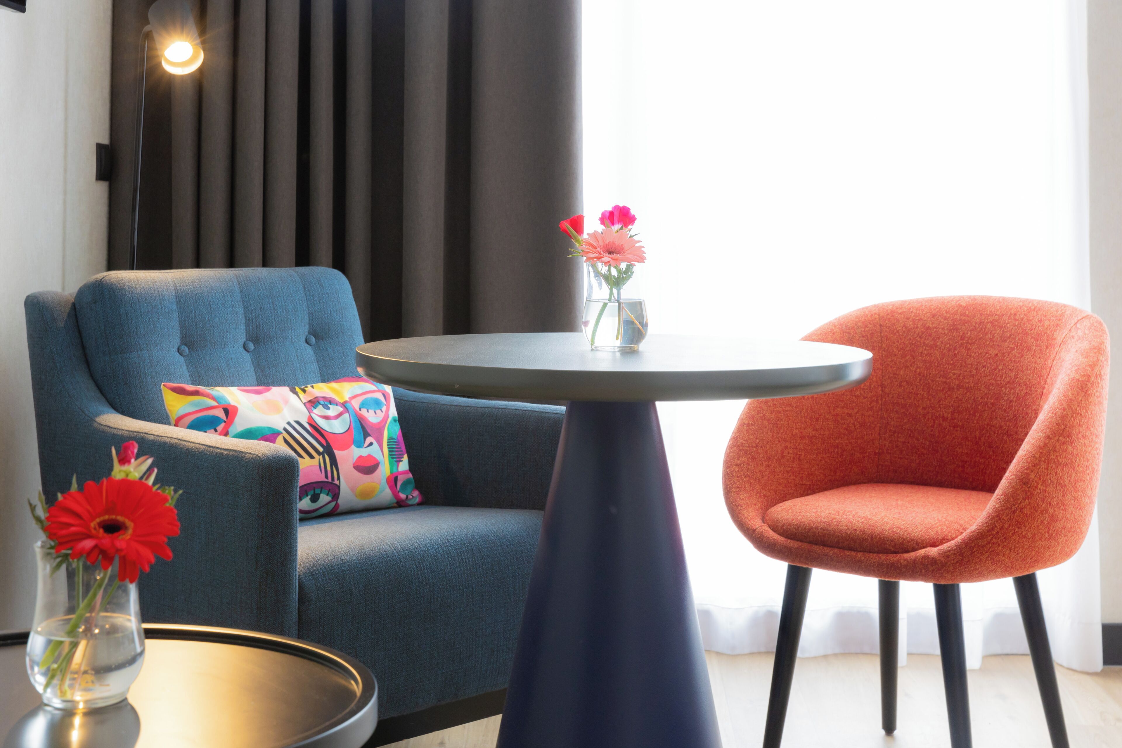 A modern living room with a blue armchair, an orange armchair, a round table, and a vase with flowers.