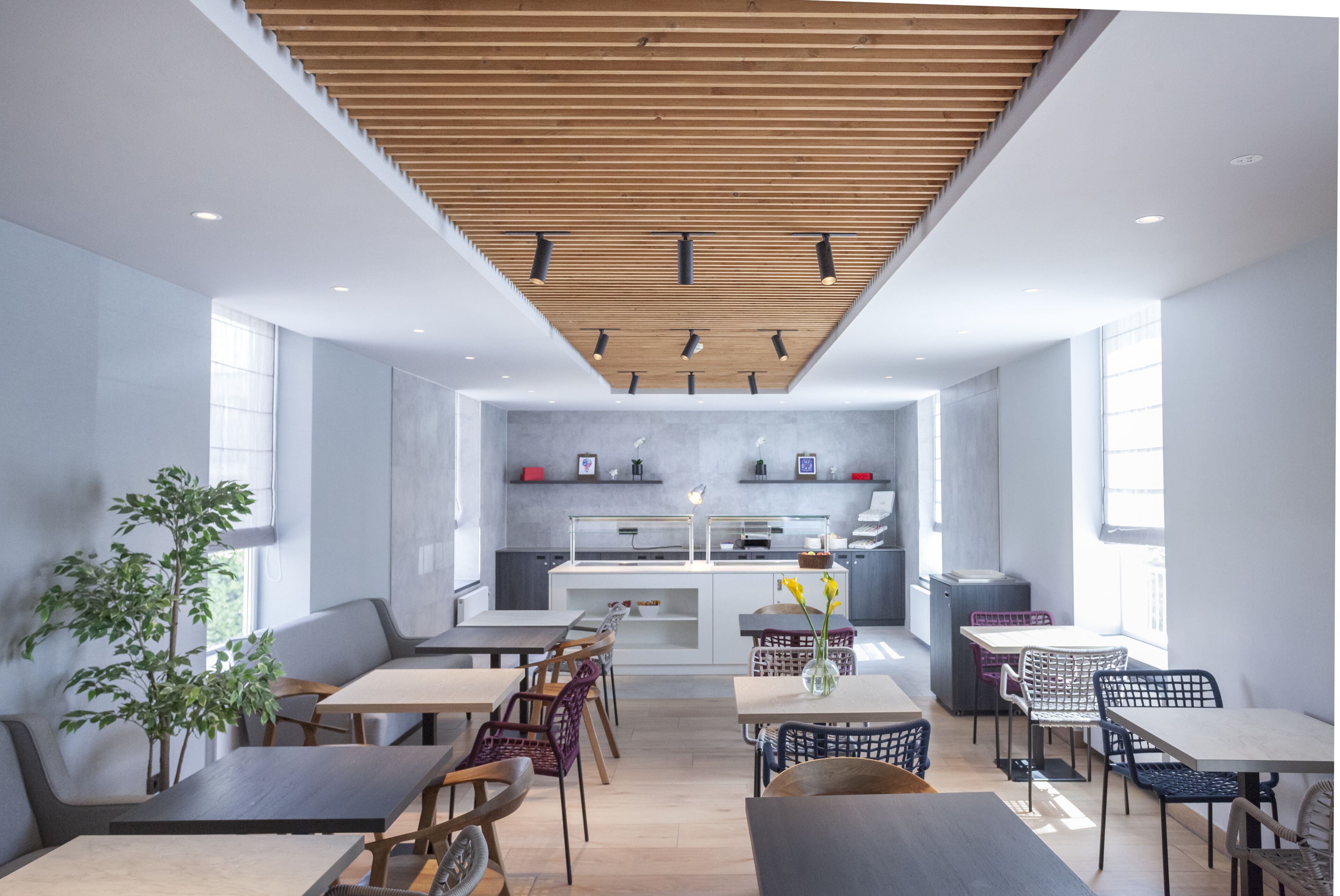 A modern dining area with wooden ceiling slats, tables, chairs, and a kitchen in the background.