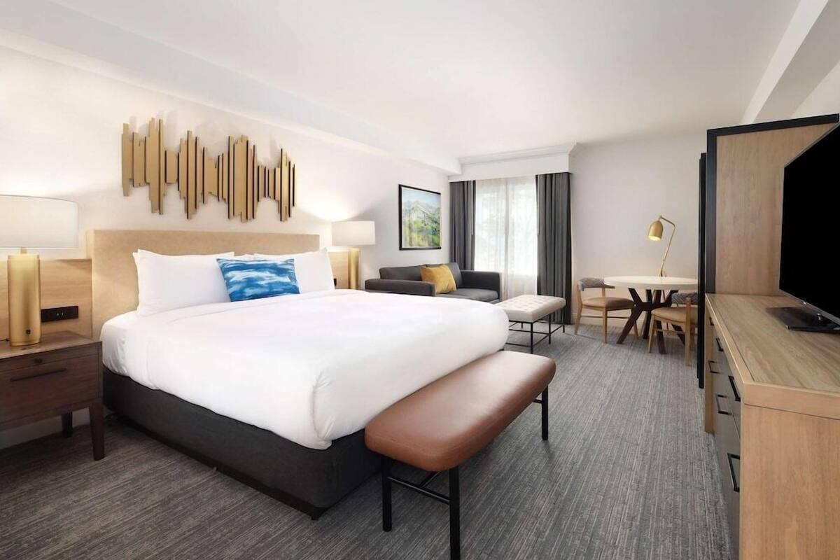 A hotel room with a large bed, a desk, a chair, a bench, a TV, and a wall art piece.