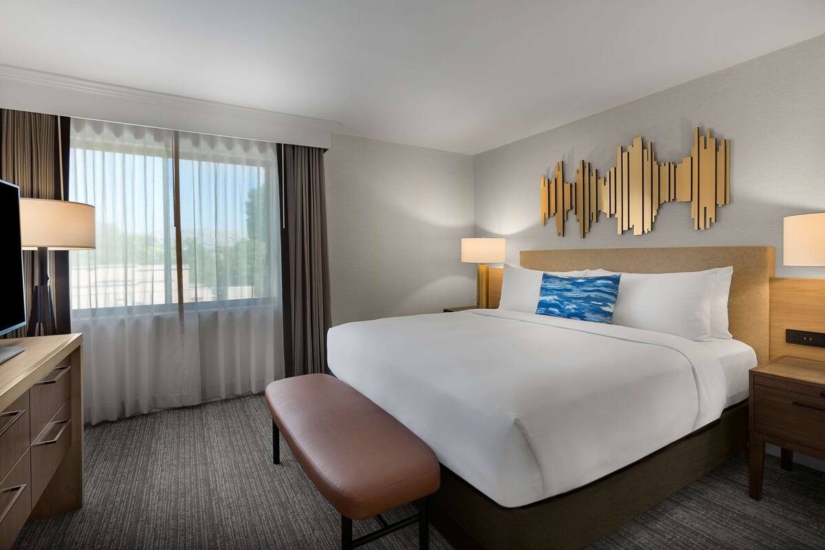A hotel room with a large bed, a desk, a bench, two bedside lamps, and a wall art piece.