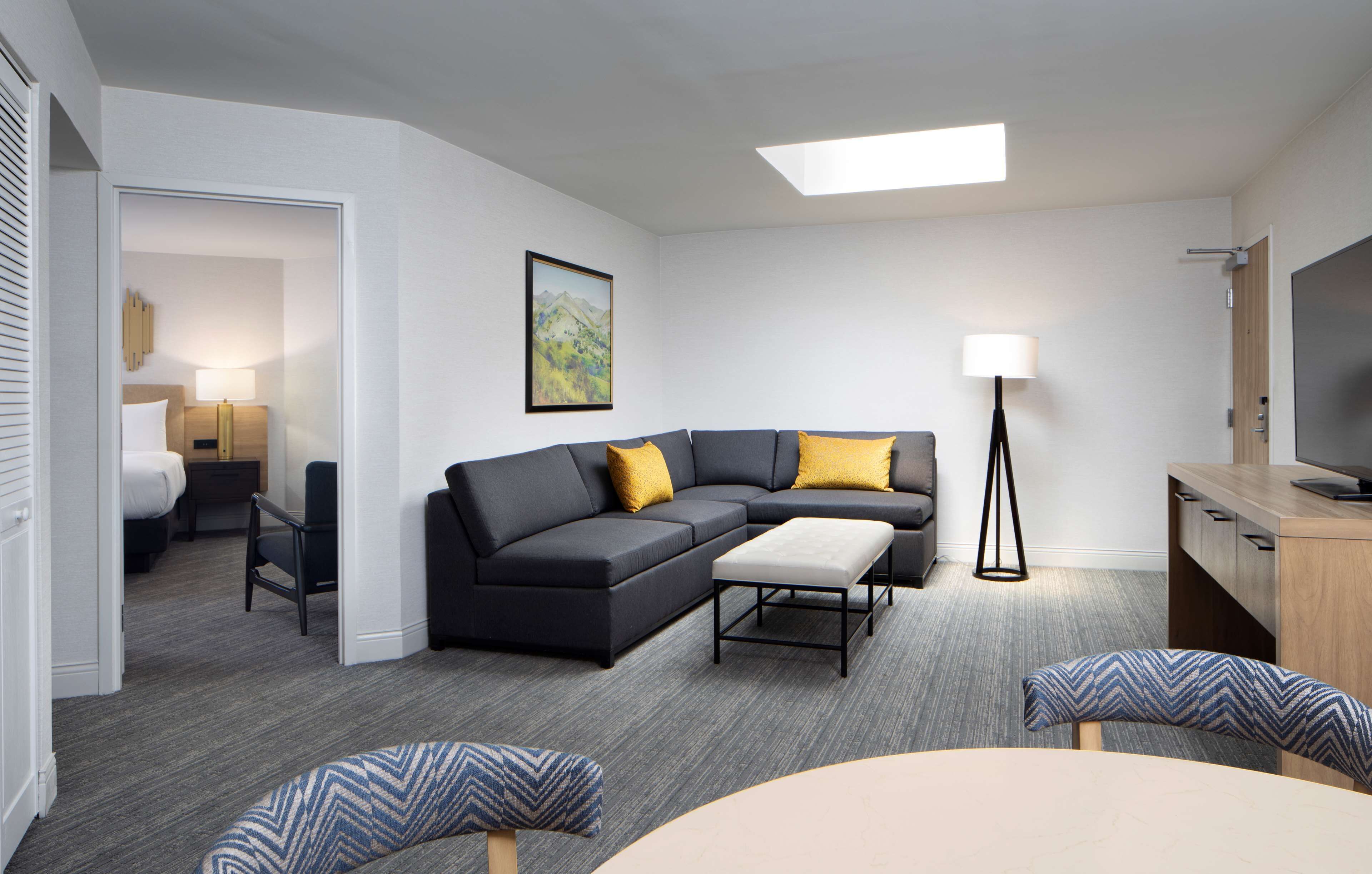 A modern hotel room with a sectional sofa, a small dining table, a desk, and a TV.