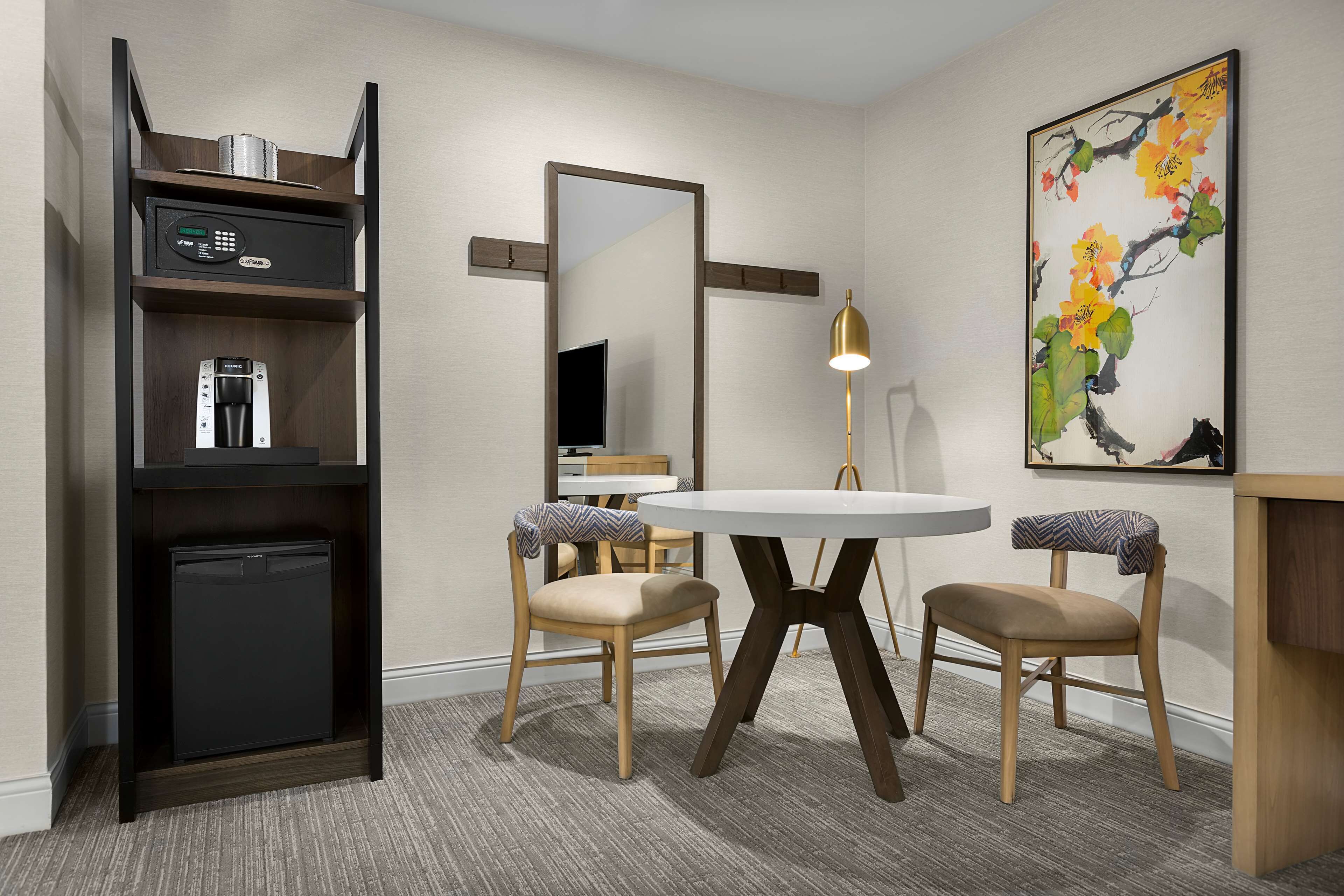 Habitación de hotel con distribución compacta, que incluye un refrigerador pequeño, cafetera, televisor, mesa redonda de comedor con dos sillas y un espejo grande.
