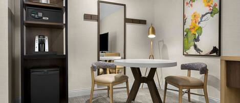 Habitación de hotel con distribución compacta, que incluye un refrigerador pequeño, cafetera, televisor, mesa redonda de comedor con dos sillas y un espejo grande.