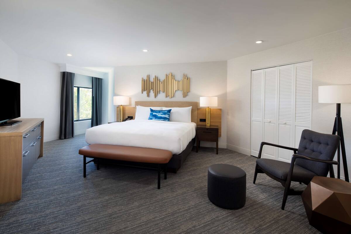 A hotel room with a bed, bedside tables, a chair, a bench, a TV, and a wardrobe.