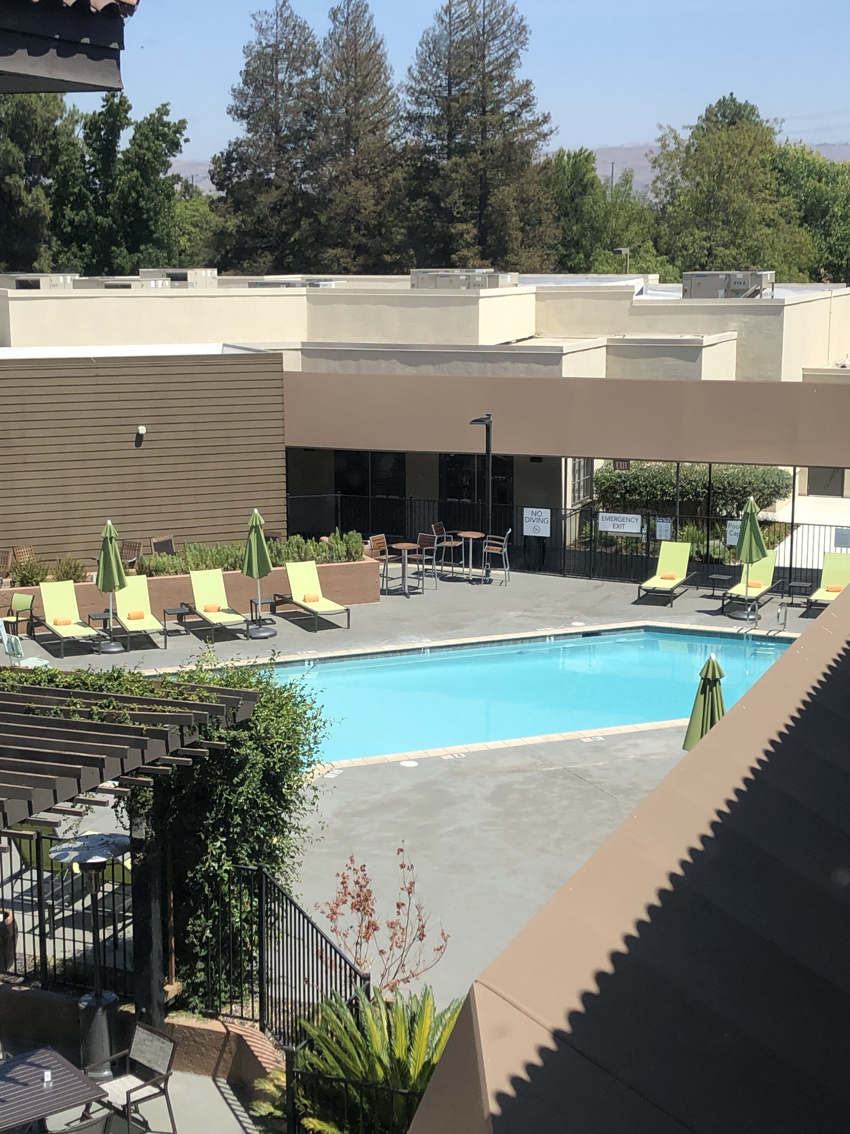 A pool area with lounge chairs and umbrellas, a sign indicating an emergency exit, and a view of trees and buildings in the background.