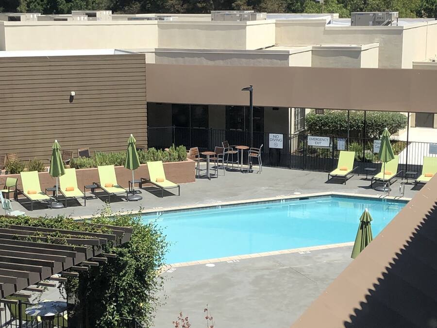 Zona de piscina con sillas de descanso y sombrillas, un letrero que indica una salida de emergencia, y vistas a árboles y edificios al fondo.