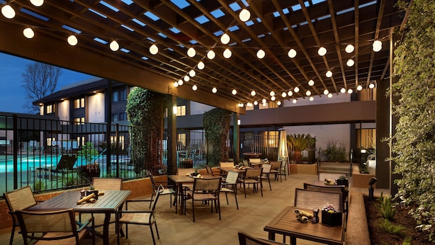 Un patio al aire libre con mesas y sillas, una piscina al fondo y una sección cubierta con techo de madera y luces colgantes.