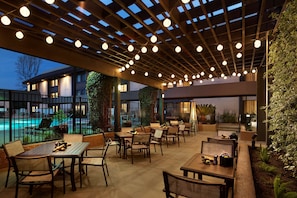 An outdoor patio area with tables and chairs, a pool in the background, and a covered section with a wooden ceiling and hanging lights. - Sonesta Silicon Valley (Milpitas)