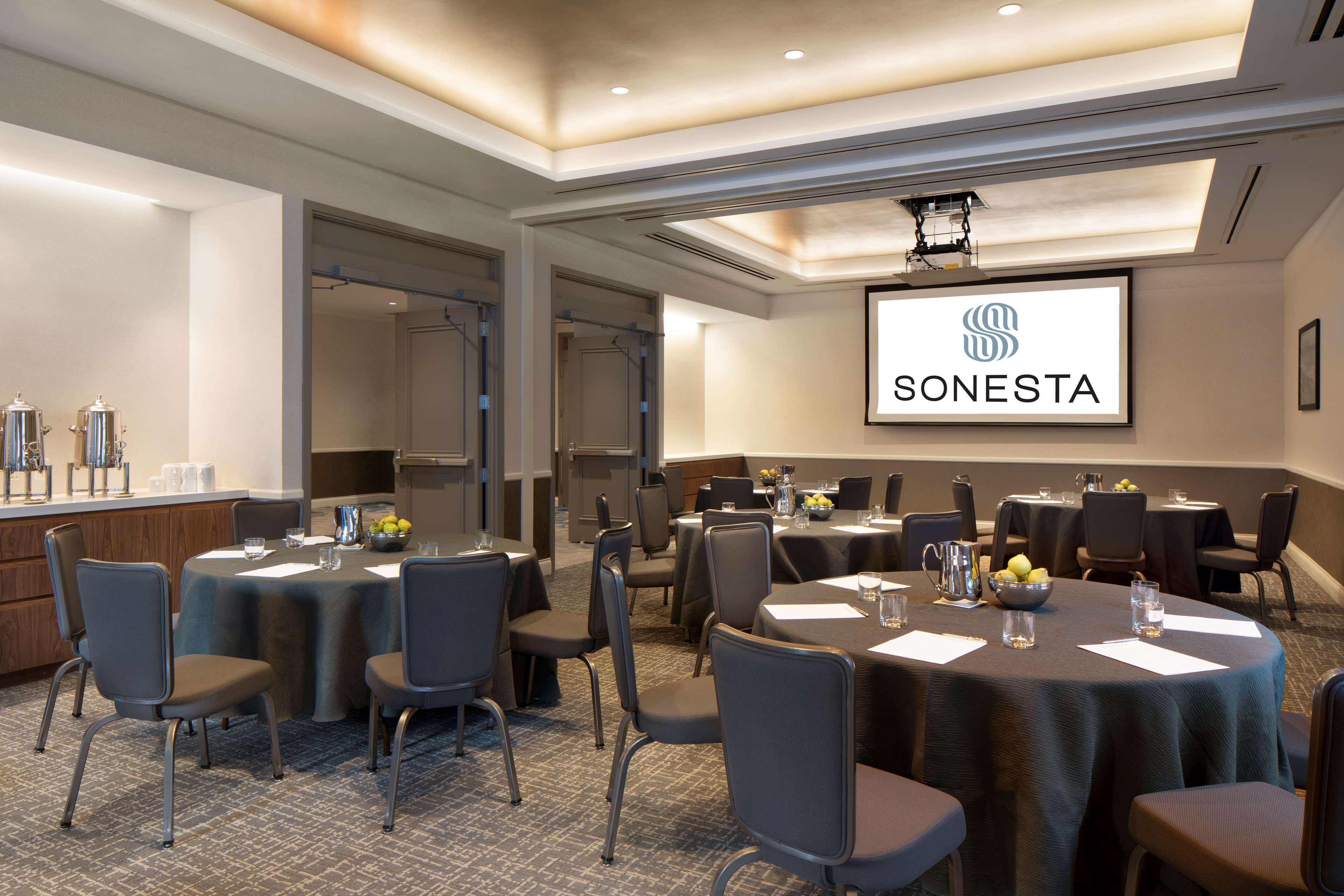 A conference room with multiple tables and chairs, a projector screen displaying the SONESTA logo, and a buffet area with food and drinks.