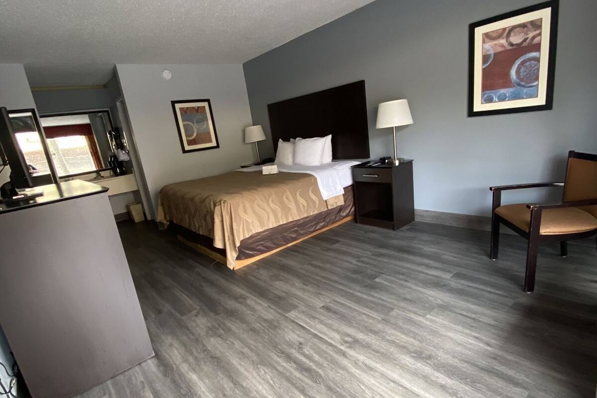 A hotel room with a bed, bedside tables, a desk, a chair, and a painting on the wall.