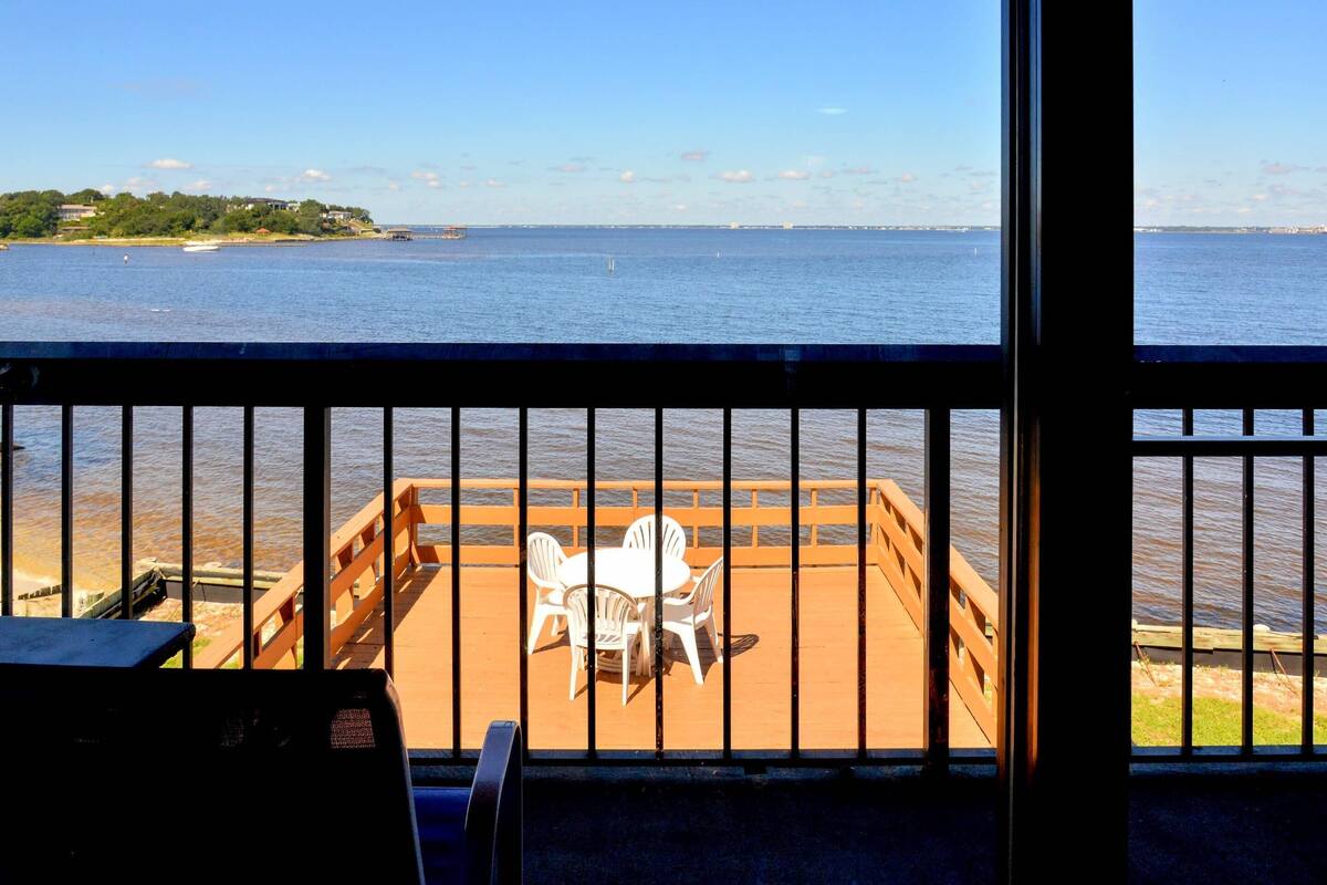 A view from a balcony with a railing, a table, and chairs facing a body of water.