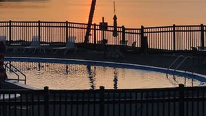 A sunset view over a calm body of water with a palm tree and a pool area with lounge chairs.