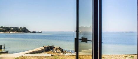 Vista desde un espacio exterior cubierto con una banca, una puerta de vidrio y el mar con la costa al fondo.