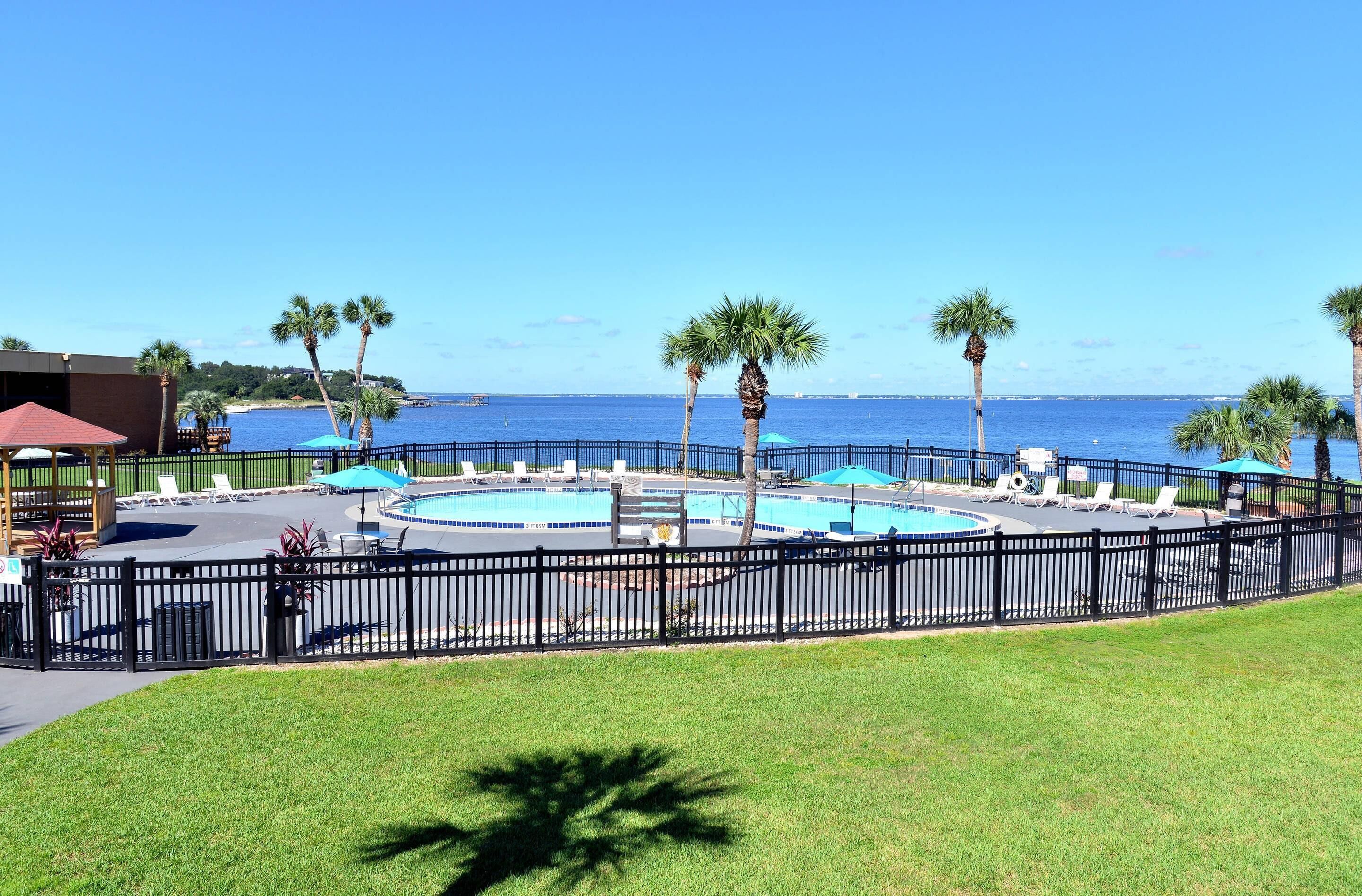 A pool area with lounge chairs and umbrellas, surrounded by a fence and palm trees, with a view of the ocean.